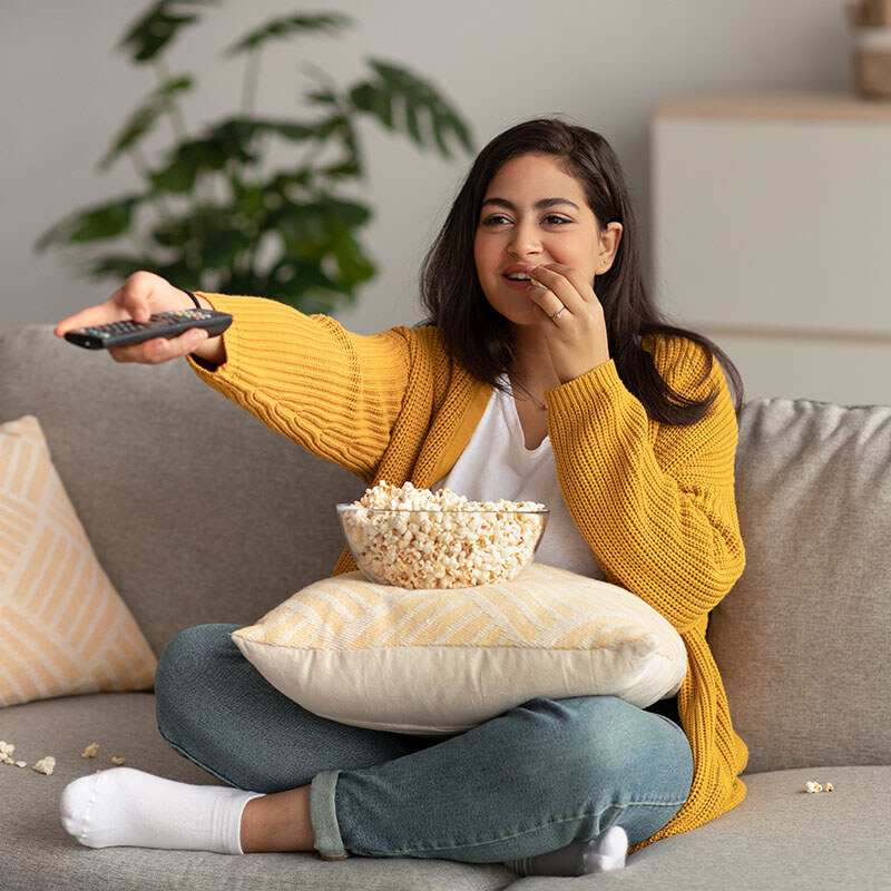 woman eating popcorn on her couch and pointing the television remote