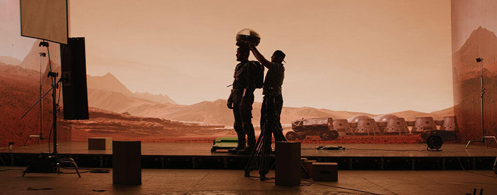 man putting helmet on space actor infront of orange Mars backdrop