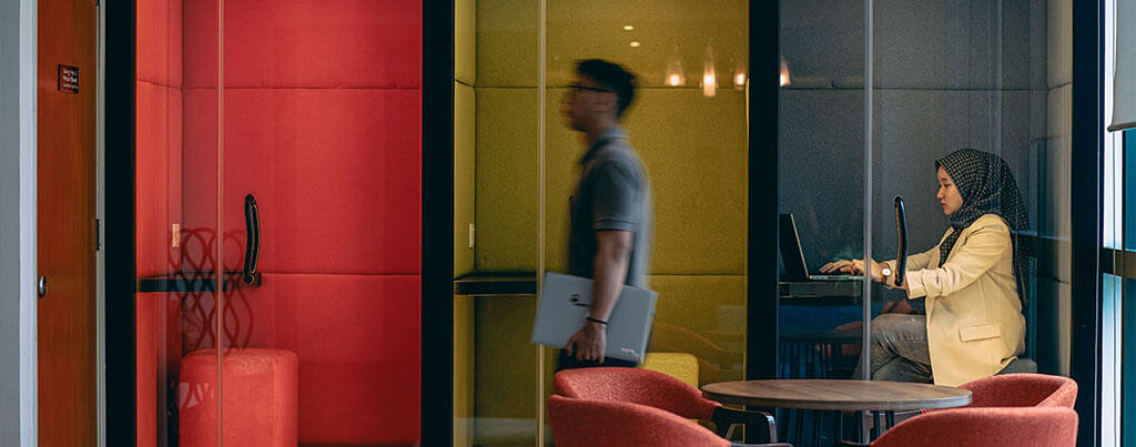 male office employee walking by privacy booth with female employee working on laptop