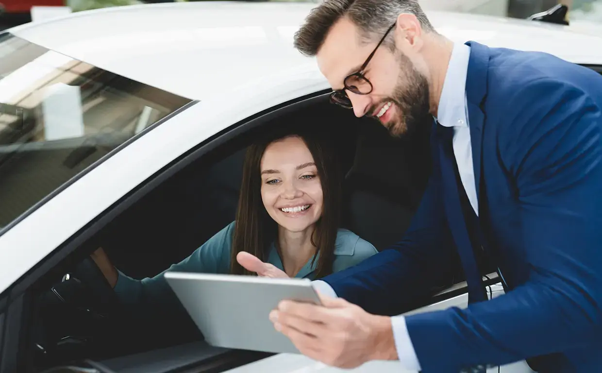 woman sitting in car and talking to sales person holding tablet outside of auto