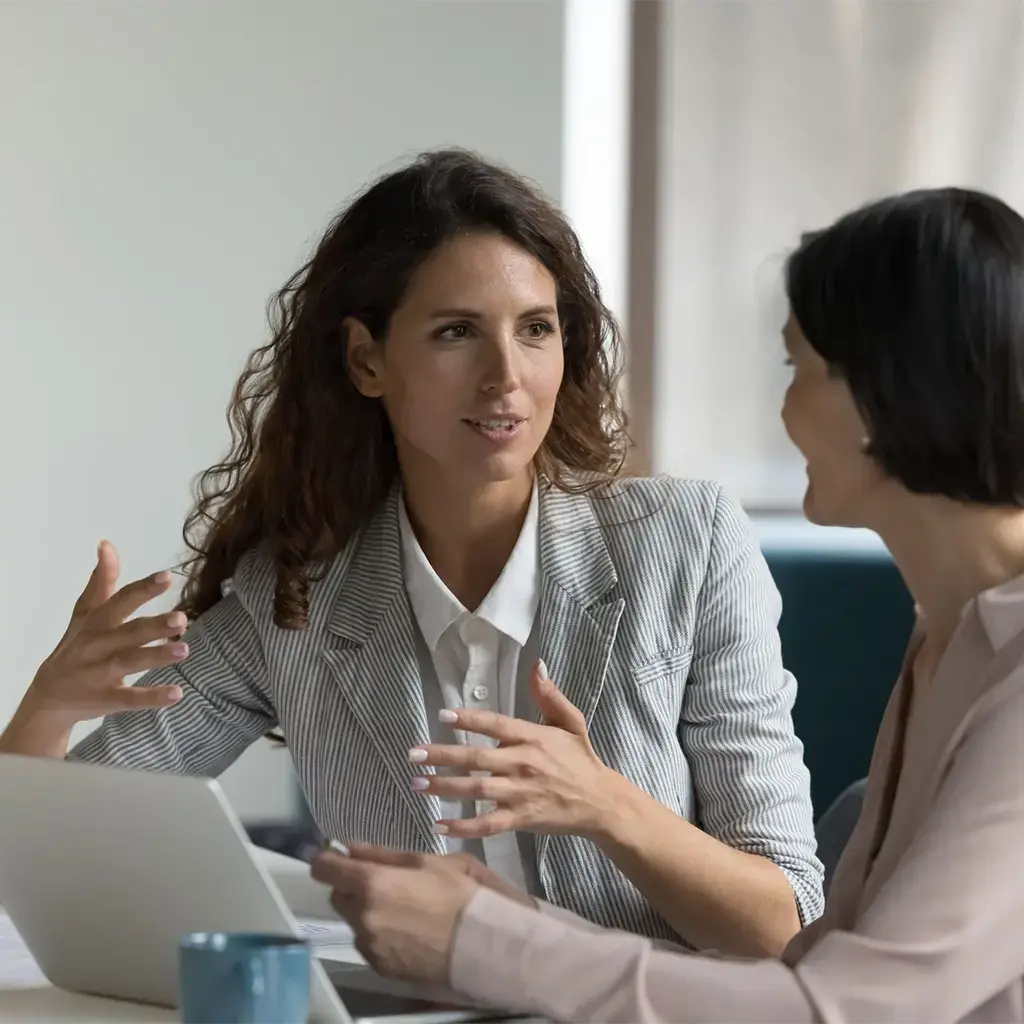 woman explaining herself using her hands and looking at another business woman working on a laptop