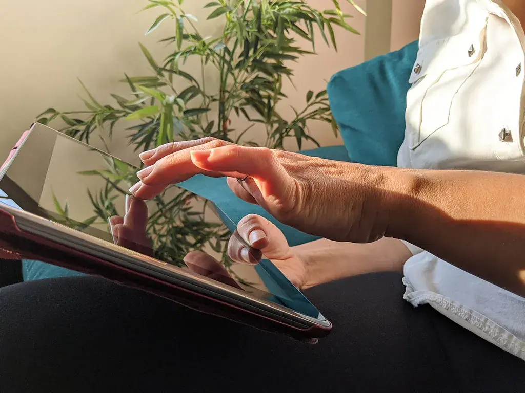 Person using a touchscreen tablet while sitting in a relaxed setting with natural light and greenery in the background.