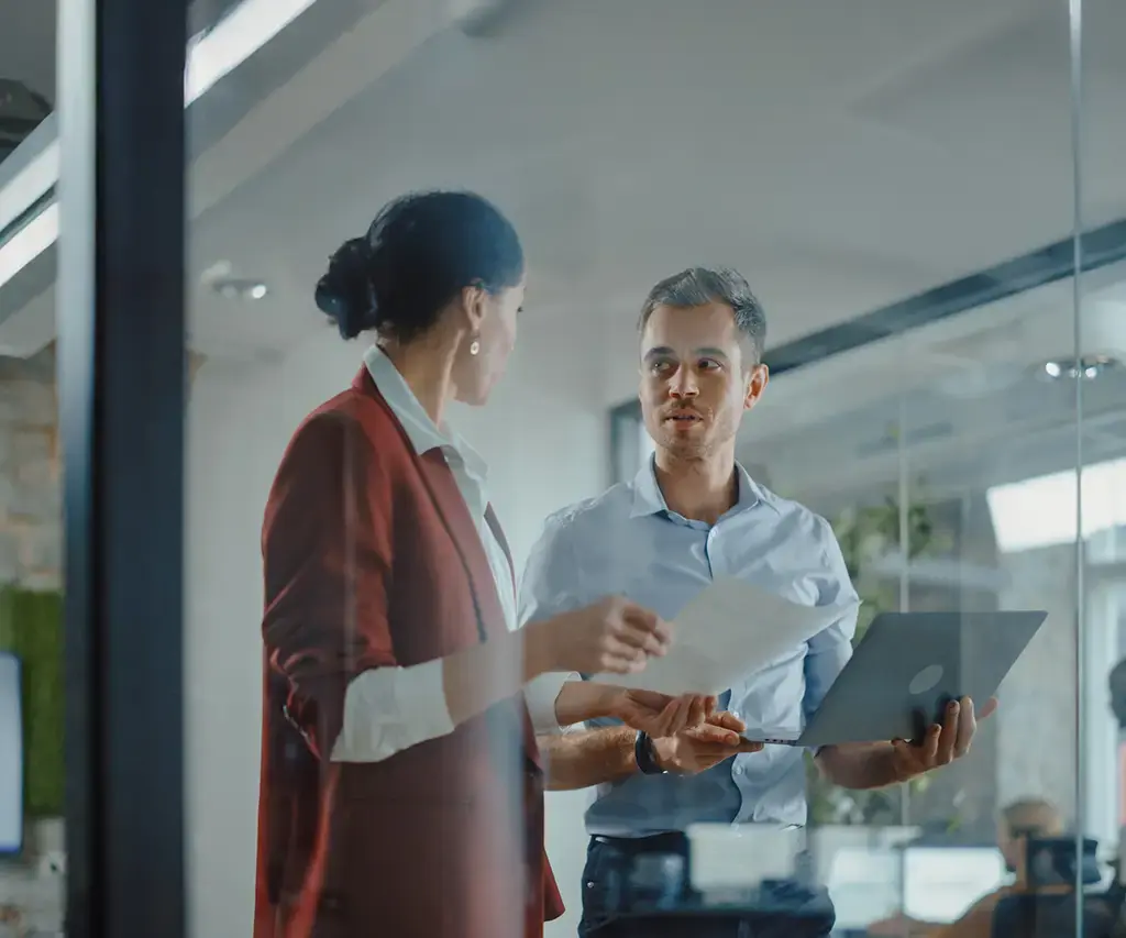 Two professionals discuss documents and a laptop in a modern office with glass walls and an open workspace.