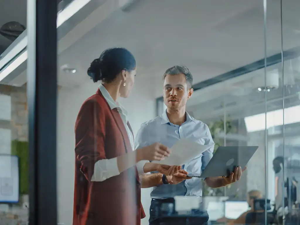 Two professionals discuss documents and a laptop in a modern office with glass walls and an open workspace.