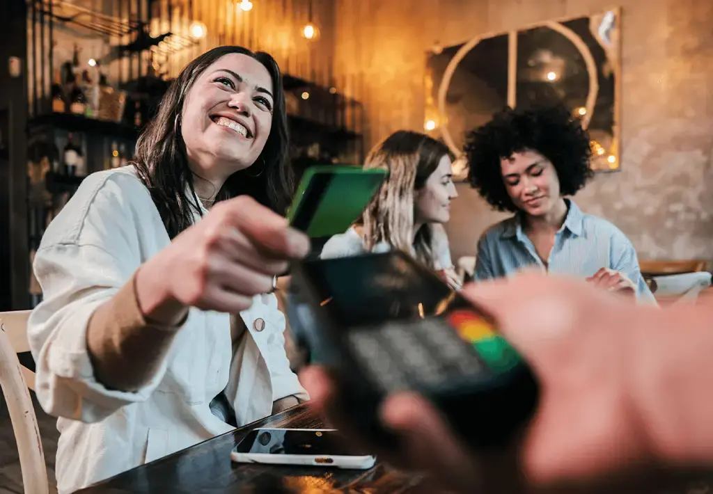 Smiling customer makes a contactless payment with a card at a restaurant while sitting with friends.