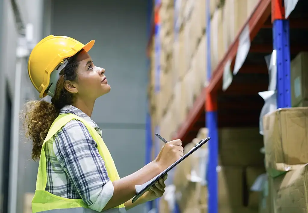 Warehouse worker in a safety vest and helmet inspecting inventory on high shelves with a clipboard.