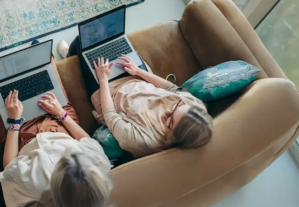 Two people sitting on a sofa, each using a laptop. They are engaged in some work or collaboration. They are looking at their screens and typing.