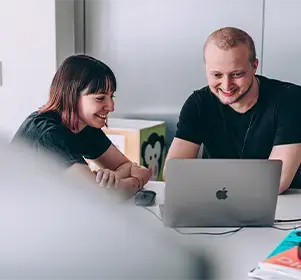 Two colleagues collaborating and smiling while working on a laptop in a modern office setting.