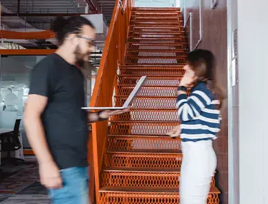 Two colleagues walking past each other in a modern office, one holding a laptop, near bright orange stairs.