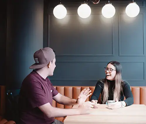 Two colleagues having a casual conversation over coffee at a wooden table in a modern office setting