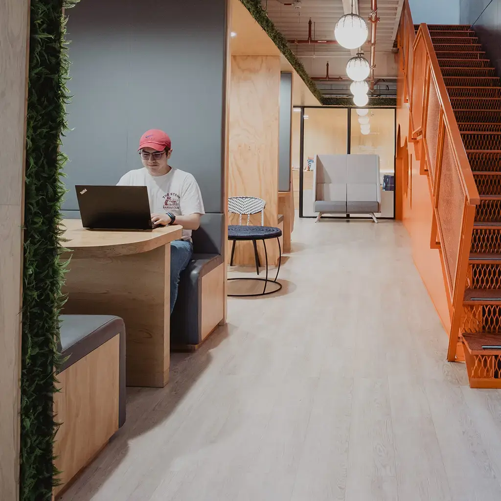 Endava employee sits in a wooden booth and he is working on a laptop near a set of orange stairs going up a floor