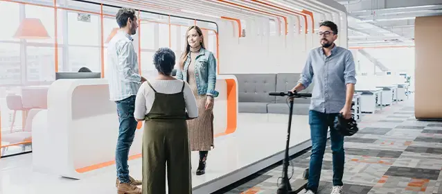 Colleagues interacting in a bright, open office lobby, with one person walking past with a scooter.