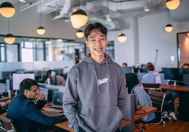 A professional smiling in a modern open office environment with colleagues working in the background.