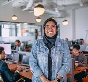 A professional smiling confidently in a modern open office environment with colleagues working in the background.