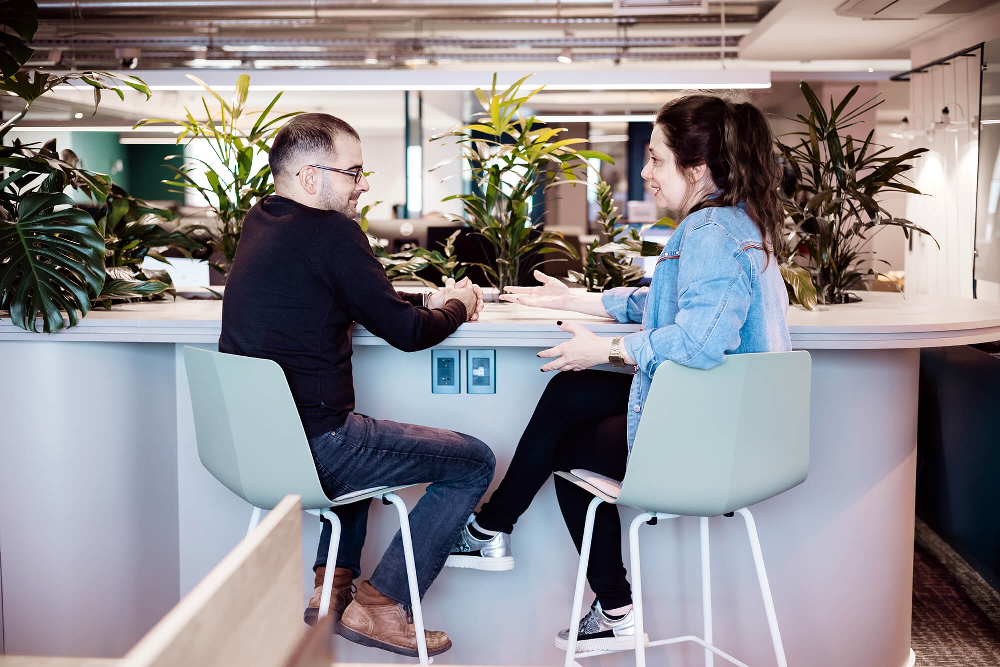 Two colleagues discussing work while seated at a modern office bar area with greenery in the background.