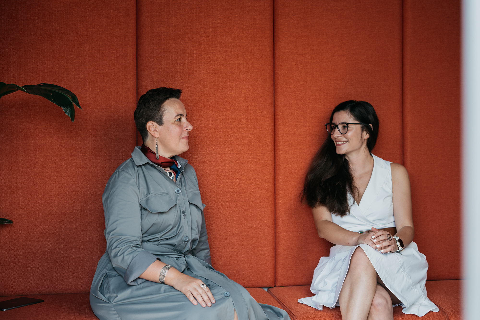 Two colleagues sitting on an orange bench in conversation, fostering collaboration in a modern office setting with an orange wall