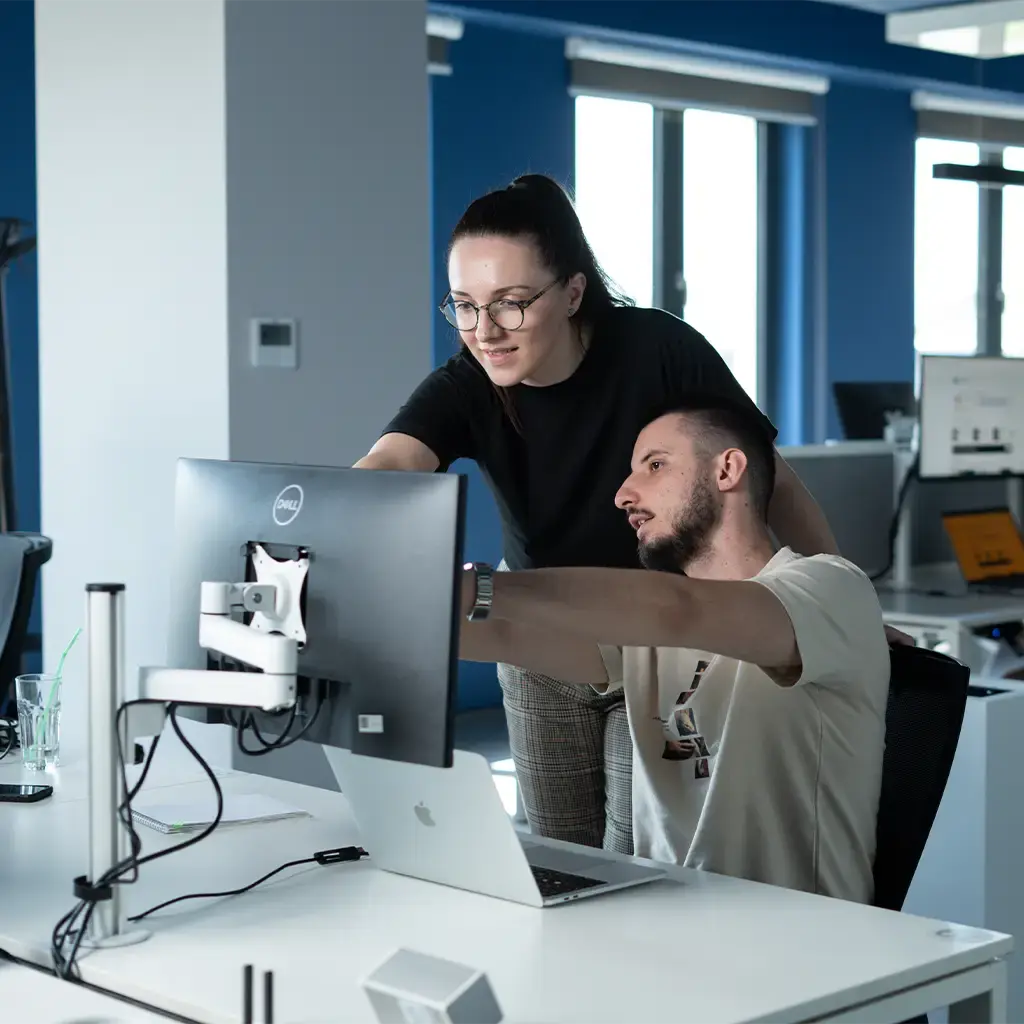 Intern collaborating with an Endava employee in a modern office, reviewing code on a monitor as they discuss a project.