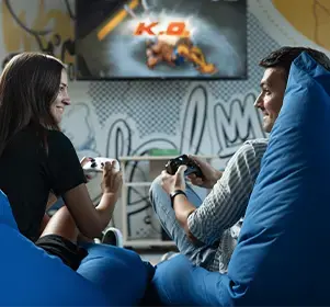 Two colleagues enjoying a video game on a screen while seated on blue bean bags in a casual office space.