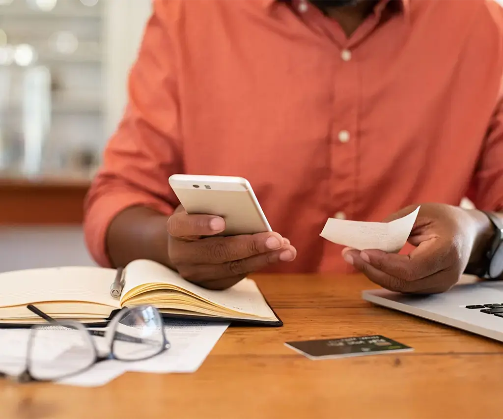 black man in orange shirt holding mobile phone near credit card and laptop 