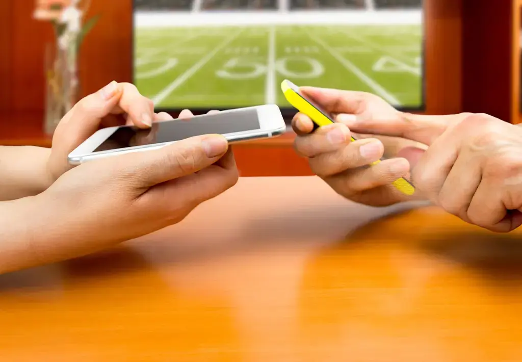 two people holding phones in front of television showing sports field with numbers