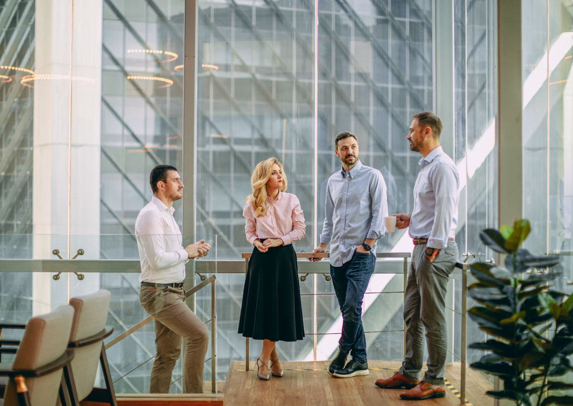 Three men in slacks and collared shirts stand with a woman wearing a long pleated skirt and a collared shirt in the corner of an office building surrounded by windows