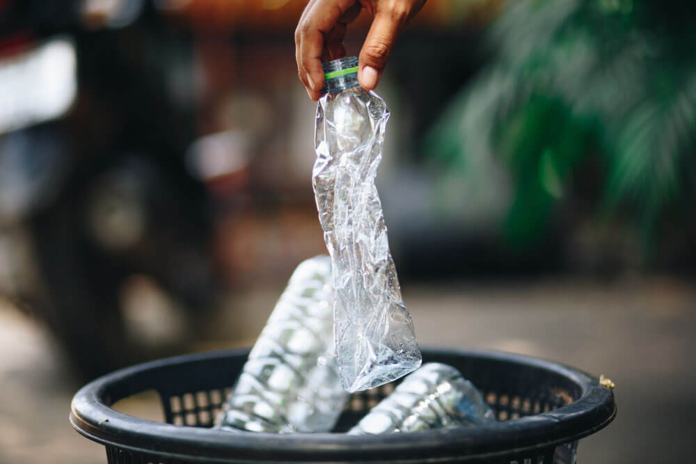 hand holding a crumpled water bottle over a recycling trash can