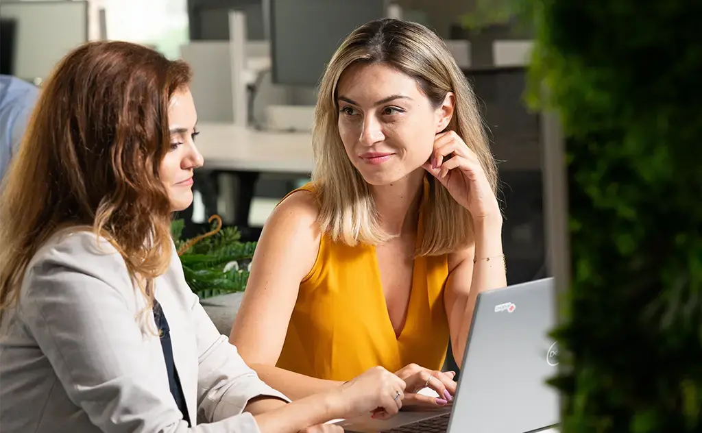 woman in dark yellow shirt looking at her coworker who is looking at her laptop